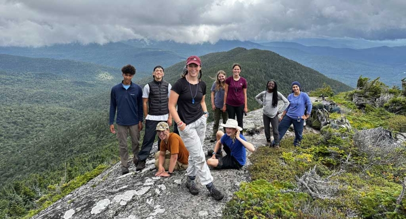 A group of people stand at the summit of a mountain, with green mountains continuing in the distance. 